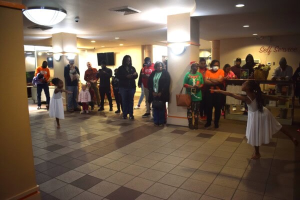 Two young girls in white dresses perform a dance in a lobby as a diverse group of adults and children watch. The audience stands in a semi-circle, with some people taking photos. The atmosphere appears warm and attentive.