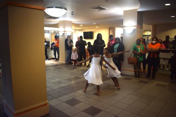 Two young girls in white dresses dance in the center of a tiled room, while a group of adults and children watch them from the background, some smiling and taking photos. The room is warmly lit with columns and ceiling lights.