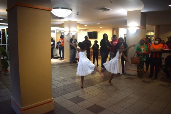 Two young girls in white dresses perform a dance in a lobby area, while a group of people watch and stand around them. The setting is warmly lit with tiled floors and light-colored pillars.