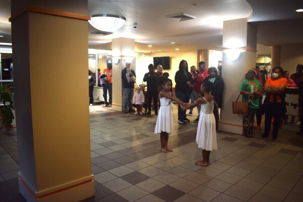 Two young girls in white dresses hold hands and perform a dance in a lobby, while a group of adults and children stand around and watch. The area is warmly lit and the audience appears engaged.