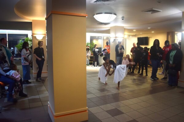 Two young dancers in white costumes perform in a lobby area as a diverse audience stands and watches. The space is well-lit with tiled floors and beige pillars, and people are gathered around attentively.