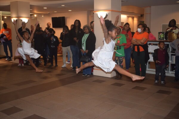 Two young girls in white dresses leap gracefully in a dance performance indoors, while a group of people, including adults and children, watch attentively in the background.