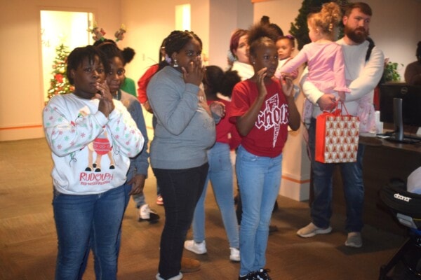 A group of children and adults stand indoors, some with hands on their faces, appearing surprised or excited. A man holds a young girl and a gift bag. Holiday decorations are visible in the background.