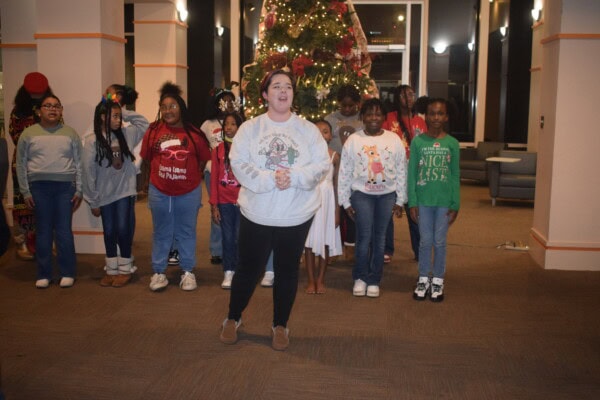 A woman stands in front of a group of children wearing festive sweaters, performing indoors near a decorated Christmas tree with lights and ornaments.