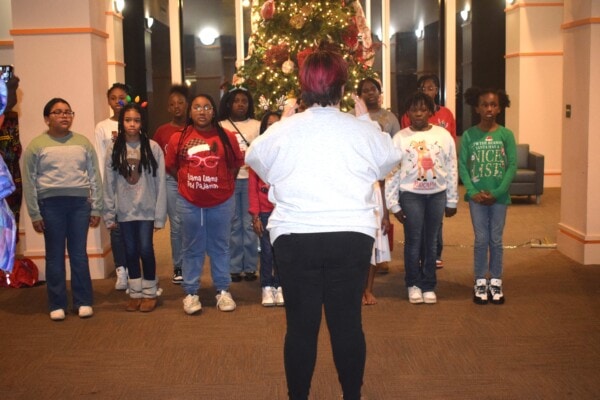 A group of children stands in rows, singing in front of a decorated Christmas tree indoors, while a woman with short hair stands facing them, conducting.