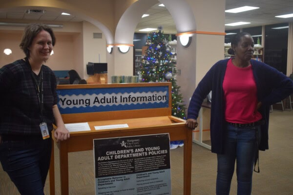 Two women stand on either side of a Young Adult Information desk in a library, smiling. A decorated Christmas tree is visible in the background.