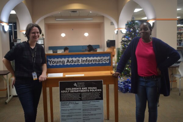 Two women stand on either side of a Young Adult Information desk in a library. Behind them, people sit at tables and a decorated Christmas tree is visible to the right. A policies sign is displayed on the desk.