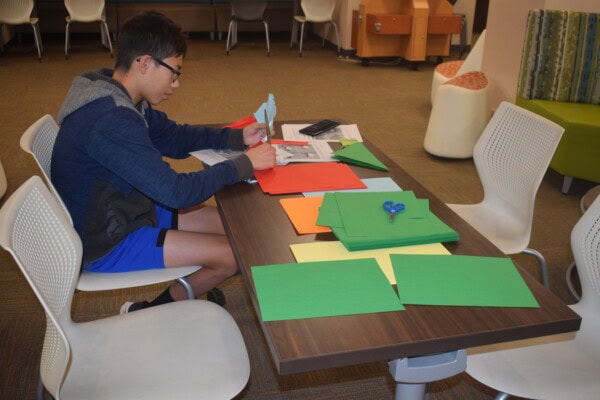 A boy sits at a table cutting blue paper, surrounded by colorful sheets, scissors, and other craft materials in a room with white chairs and a green couch.