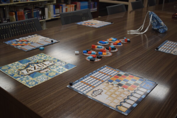 A table set up for the board game Azul, with four player boards, circular trays holding colorful tiles, a cloth tile bag, and bookshelves in the background.
