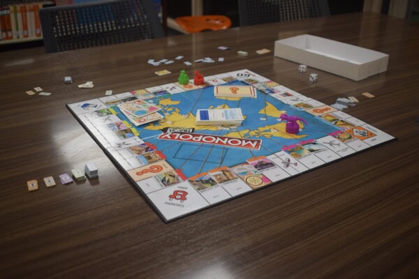 A Monopoly board game is set up on a wooden table, with cards, dice, game pieces, and Monopoly money scattered around. The game box lid sits to the side, and empty chairs surround the table.