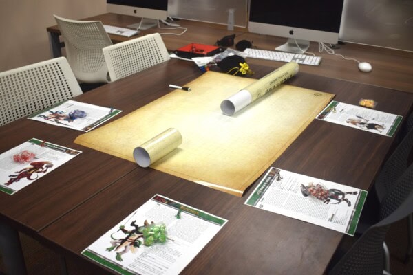 A tabletop gaming setup with a blank grid map, dice, character sheets, and miniatures at each seat, surrounded by white chairs in a room with desktop computers in the background.