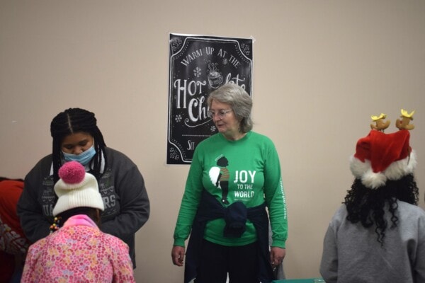 A woman in a green Joy to the World shirt stands by a Hot Chocolate sign, surrounded by children in festive winter hats and a mask, at what appears to be a holiday event.
