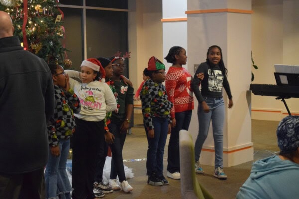 A group of children wearing festive holiday clothing stand in a line indoors near a decorated Christmas tree, singing or performing together. A digital piano and a few adults are visible in the background.