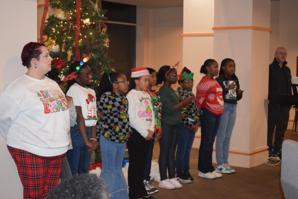 A group of children dressed in festive holiday sweaters and accessories stand in a line, singing in front of a decorated Christmas tree. An adult and a keyboard player are nearby.