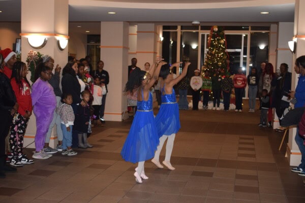 Two young girls in blue dresses perform a ballet dance in the center of a room, surrounded by an audience. A decorated Christmas tree stands in the background, and the audience watches attentively.