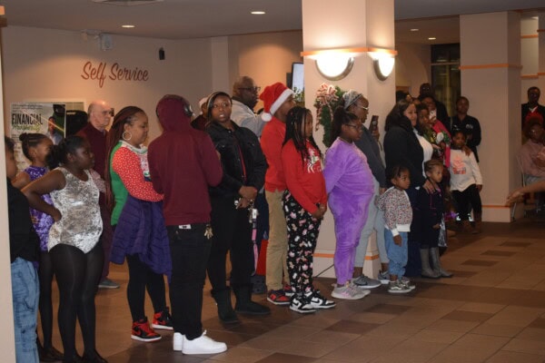 A group of people, including children and adults, stand in a line indoors. Some wear festive clothing and Santa hats, while others are casually dressed. The atmosphere appears cheerful and social.