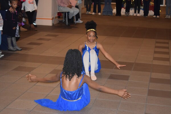 Two young girls in blue dresses are performing a dance routine on a tiled floor indoors, sitting on the ground facing each other with arms outstretched, while a small crowd watches in the background.