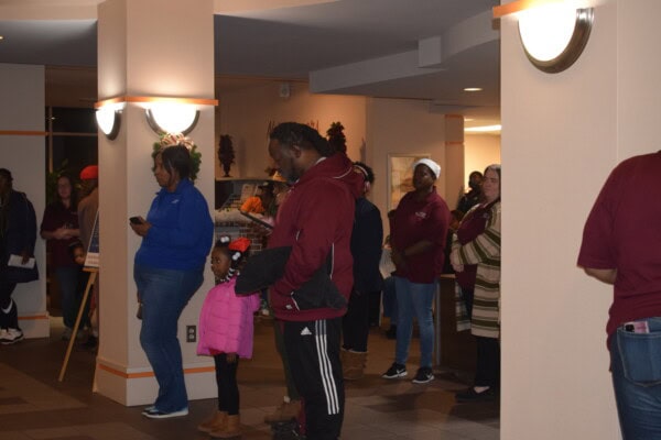 A group of people, including adults and children, stand in a well-lit lobby. Some are looking at their phones, while others wait in line or talk quietly. One child in a pink jacket stands near an adult in maroon clothing.