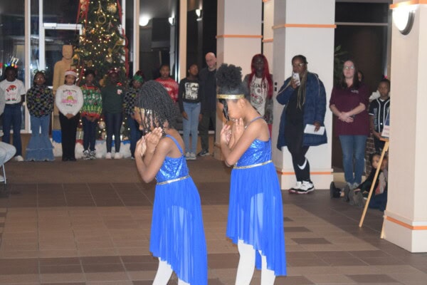 Two young girls in matching blue dresses perform a dance in the foreground, while a group of people, some wearing holiday sweaters, watch near a decorated Christmas tree in the background.