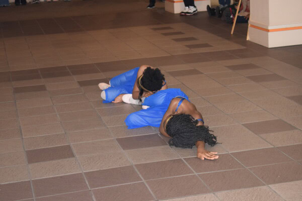 Two dancers in blue costumes lie on a tiled floor, performing a contemporary dance routine. One dancer is lying on her side with her arm extended, while the other is curled up nearby. Spectators are partially visible in the background.