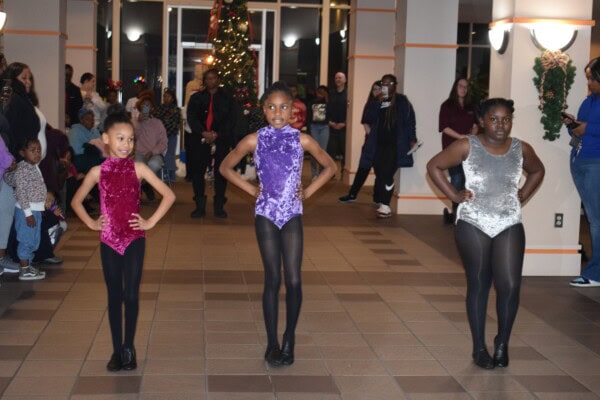 Three young girls in sparkly dance costumes stand in a row, posing with hands on hips. People watch from the background, and a decorated Christmas tree is visible behind them. The setting appears to be an indoor event.