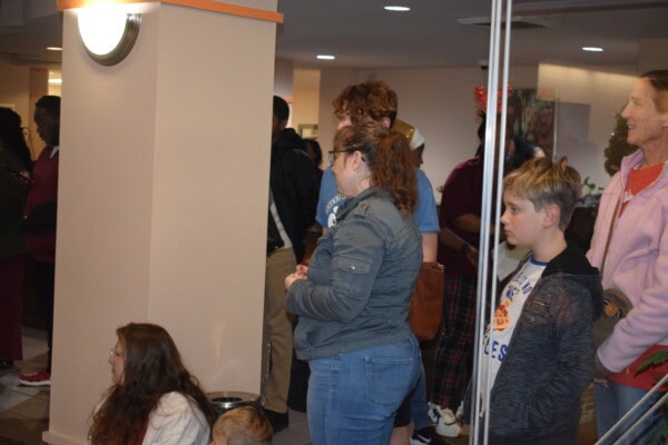 A group of people, including adults and children, stand and sit in a lobby area. Some are facing forward, others are turned to the side. The setting appears to be indoors with neutral walls and bright lighting.