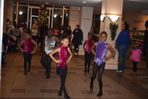 A group of young girls in sparkly dance costumes perform a routine indoors. People watch from the background, and a decorated Christmas tree is visible by the windows.