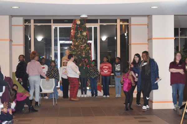 A group of people, including children and adults, gather indoors in front of a decorated Christmas tree. Some wear festive sweaters and pajamas, while others stand or sit, casually interacting in a cheerful atmosphere.