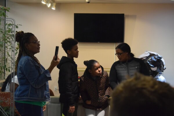 Four people stand indoors near a blank TV screen. One woman holds up a phone, while two children and another woman look at each other, appearing engaged in conversation. A plant and some bags are visible in the background.