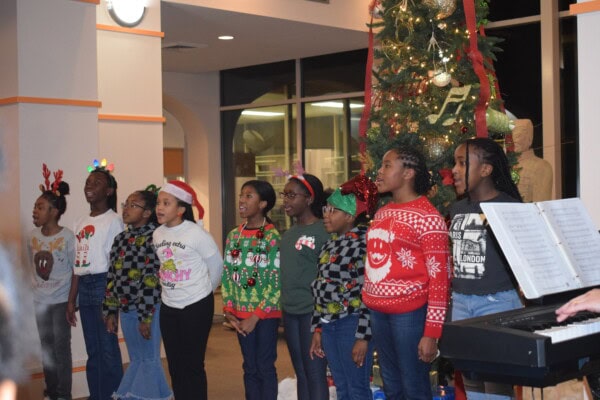 A group of children in festive sweaters and holiday accessories sing together beside a decorated Christmas tree while someone plays the keyboard nearby.