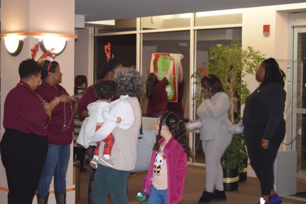 A group of adults and children, some wearing festive antler headbands, gather indoors near plants and holiday decorations. One adult holds a child, while others chat and play in a warmly lit room.