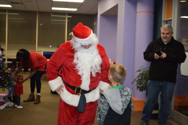 Santa Claus talks to a young boy while a woman helps two small children near a decorated Christmas tree. Another man stands nearby, looking at his phone. The scene takes place indoors in a brightly lit room.
