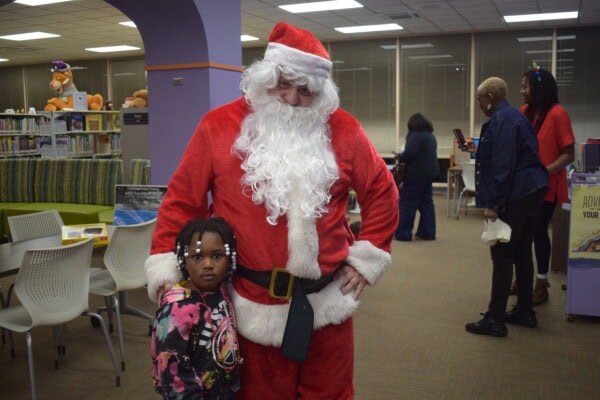 A person dressed as Santa Claus stands with a young child in a library, while several adults are in the background. The child has braided hair with beads and looks towards the camera.