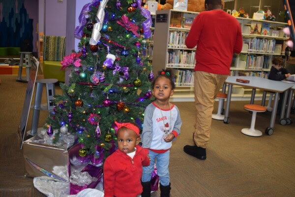 Two young children stand by a decorated Christmas tree in a library. One child wears a red outfit with a bow, while the other wears a white sweater with Santa on it. Bookshelves and two adults are visible in the background.