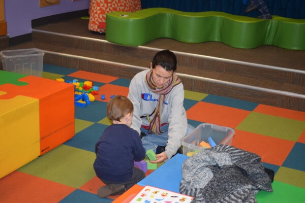 An adult and a child sit on a colorful tiled floor, playing with plastic blocks. Nearby are bins of toys, a large yellow block, and a gray sweater. The background includes green seating and scattered toys.