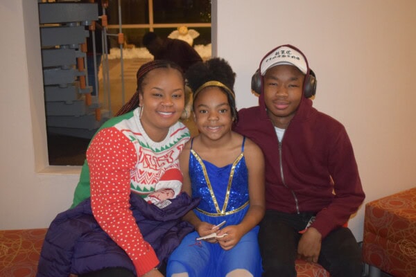 Three smiling children sit together on an orange couch. The girl on the left wears a festive sweater, the girl in the middle wears a blue dress, and the boy on the right wears a maroon jacket and headphones.