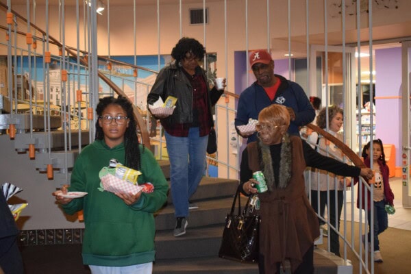 A group of people carrying food and drinks walk down a spiral staircase indoors. The group includes both adults and children, and the setting appears to be casual and social.