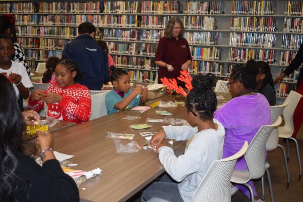 Children sit around a table in a library, working on crafts with various supplies. Bookshelves line the background, and an adult stands nearby observing the activity.