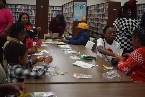 Children and adults sit around a table in a library, making crafts with colorful materials. Bookshelves line the background and a “Gift Wrapping Station” sign is visible on the wall.