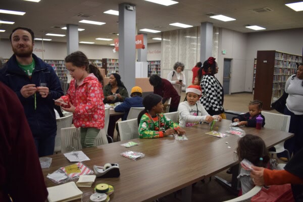 Children and adults are gathered around tables in a library, making crafts. Some children wear festive clothing. Bookshelves line the walls, and people are chatting and working together in a busy, cheerful atmosphere.