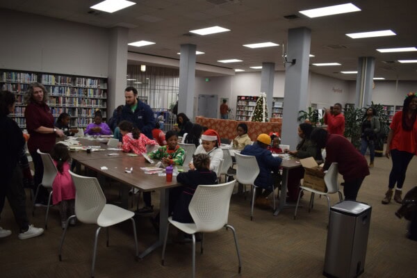 Children and adults sit around tables in a library, working on crafts together. Some people wear festive hats and sweaters. Bookshelves line the walls, and a Christmas tree is visible in the background.