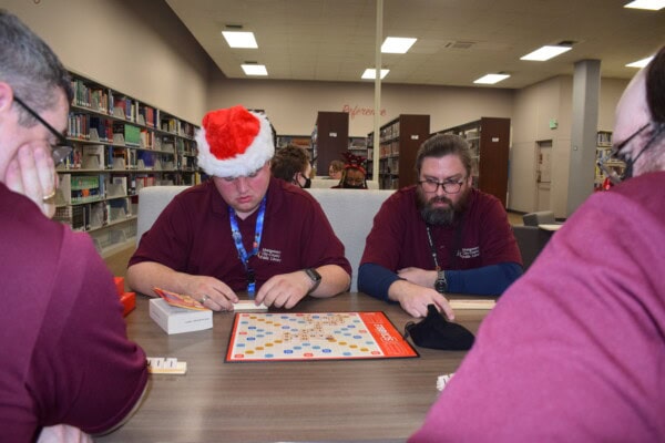 Four people in maroon shirts play Scrabble at a table in a library. One man wears a Santa hat and is focused on the board, while others watch and consider their moves. Bookshelves fill the background.