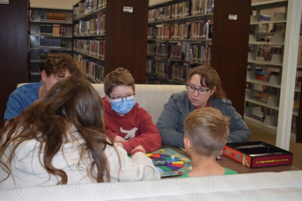 A group of five people, including children and an adult, sit around a table in a library playing a colorful board game. One child is wearing a face mask. Bookshelves are visible in the background.
