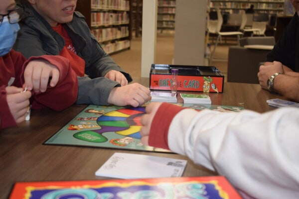 Four people sit around a table playing a board game called Queer the Card in a library. Game pieces, cards, and a timer are visible, with shelves of books in the background.