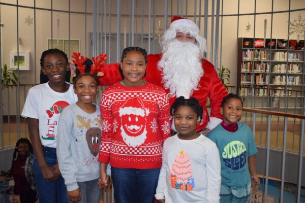 Five smiling children in festive holiday clothing stand together in front of a person dressed as Santa Claus in a library setting, with bookshelves and holiday decorations visible in the background.