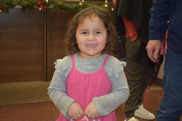 A young girl with wavy brown hair smiles at the camera. She is wearing a gray long-sleeve shirt under a pink dress. Holiday decorations with greenery and lights are in the background. An adult stands nearby.