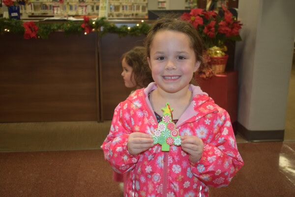 A young girl in a pink coat smiles and holds a decorated Christmas tree cookie. Another child is in the background. Holiday decorations and poinsettias are visible behind them.