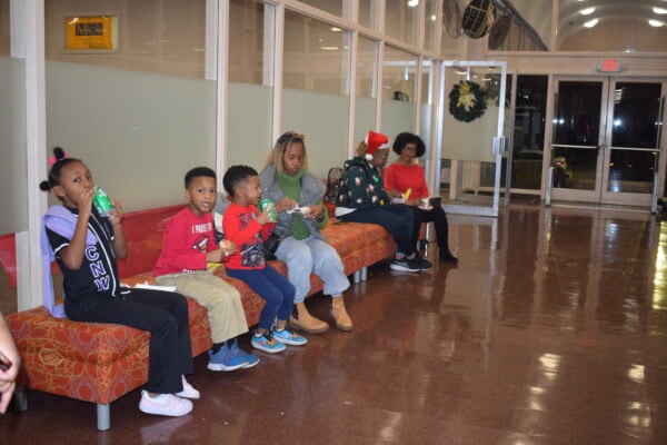 A group of children and adults sit on benches in a brightly lit hallway, drinking from juice boxes and eating snacks. Some wear festive clothing, including a Santa hat and Christmas sweater.