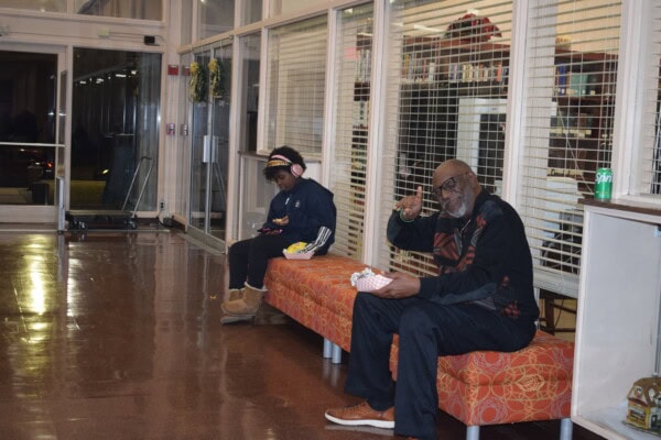 A man and a child sit on an orange patterned bench indoors. The man is smiling and making a peace sign while holding food; the child wears ear muffs and boots, looking down at a device. A closed shop is behind them.