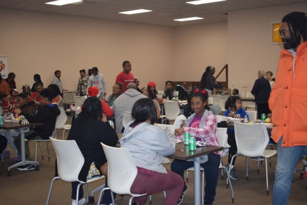 A group of people, including children and adults, sit at tables and interact in a large, well-lit room. Some people are eating or chatting, and a few are standing or walking around. The atmosphere appears lively and social.